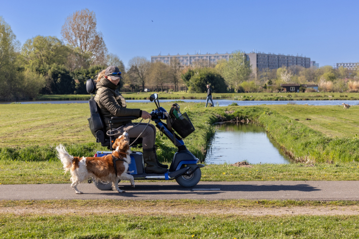 Een vrouw op een scootmobiel rijdt in het Noorderpark. Naast haar wandelt een hond mee.