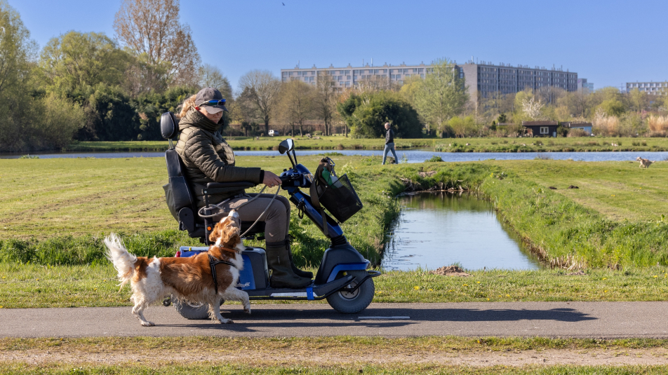 Een vrouw op een scootmobiel rijdt in het Noorderpark. Naast haar wandelt een hond mee.