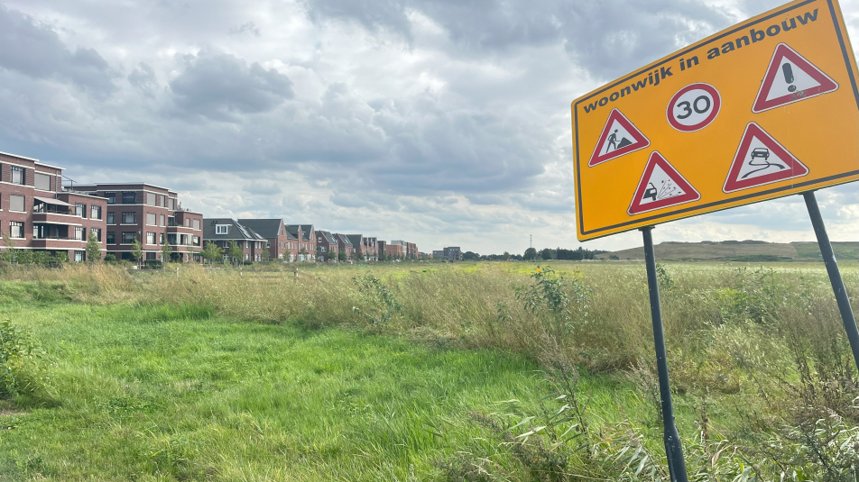 Op de voorgrond een bord met de waarschuwing 'wijk in aanbouw', op de achtergrond een leeg weiland met ernaast een woonwijk